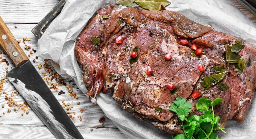 A slab of marinated beef is laid out on parchment paper, garnished with pomegranate seeds, herbs, and spices. A large knife rests beside it on a white wooden surface.