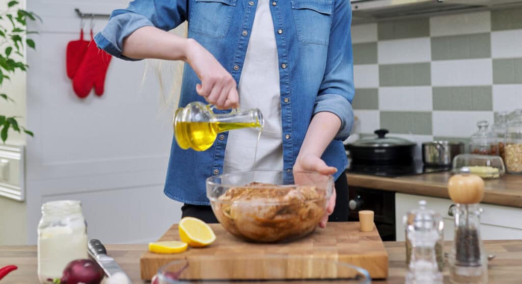 A person in a denim shirt pours olive oil into a glass bowl containing marinated chicken in a kitchen.