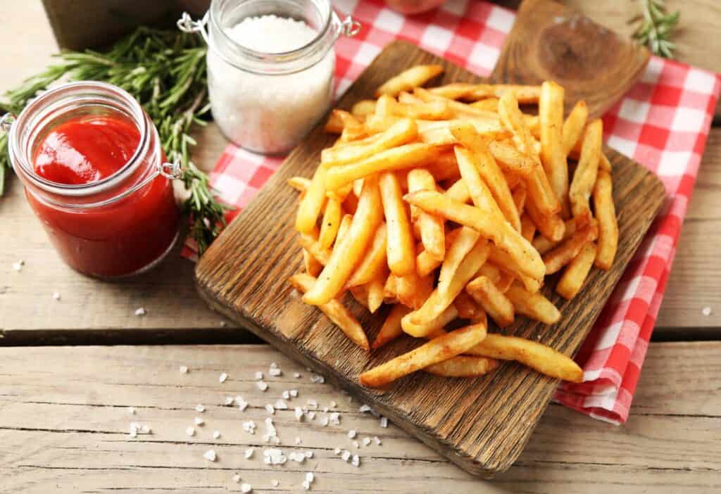 A wooden plate of seasoned French fries next to jars of ketchup and salt on a red and white checkered cloth with fresh rosemary sprigs.