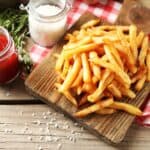 A wooden plate of seasoned French fries next to jars of ketchup and salt on a red and white checkered cloth with fresh rosemary sprigs.
