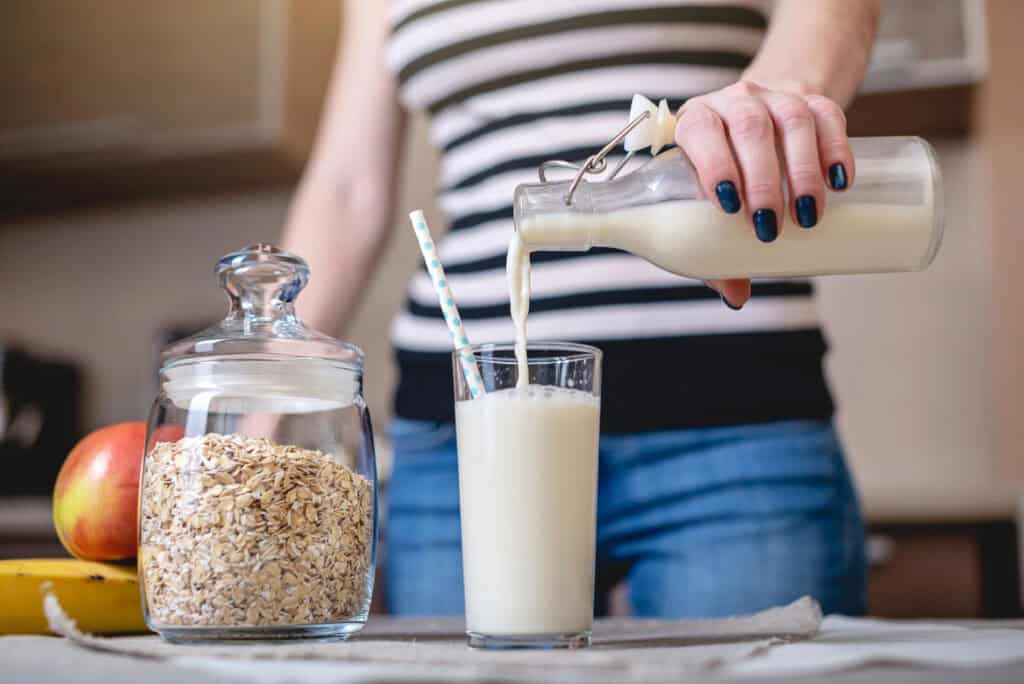 A person pours milk from a glass bottle into a tall glass beside a jar of oats, an apple, and a banana on a kitchen counter.