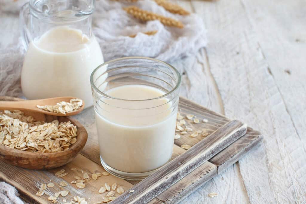 A glass and a small pitcher of oat milk are on a wooden surface. Oats and a wooden spoon filled with oats are beside them.