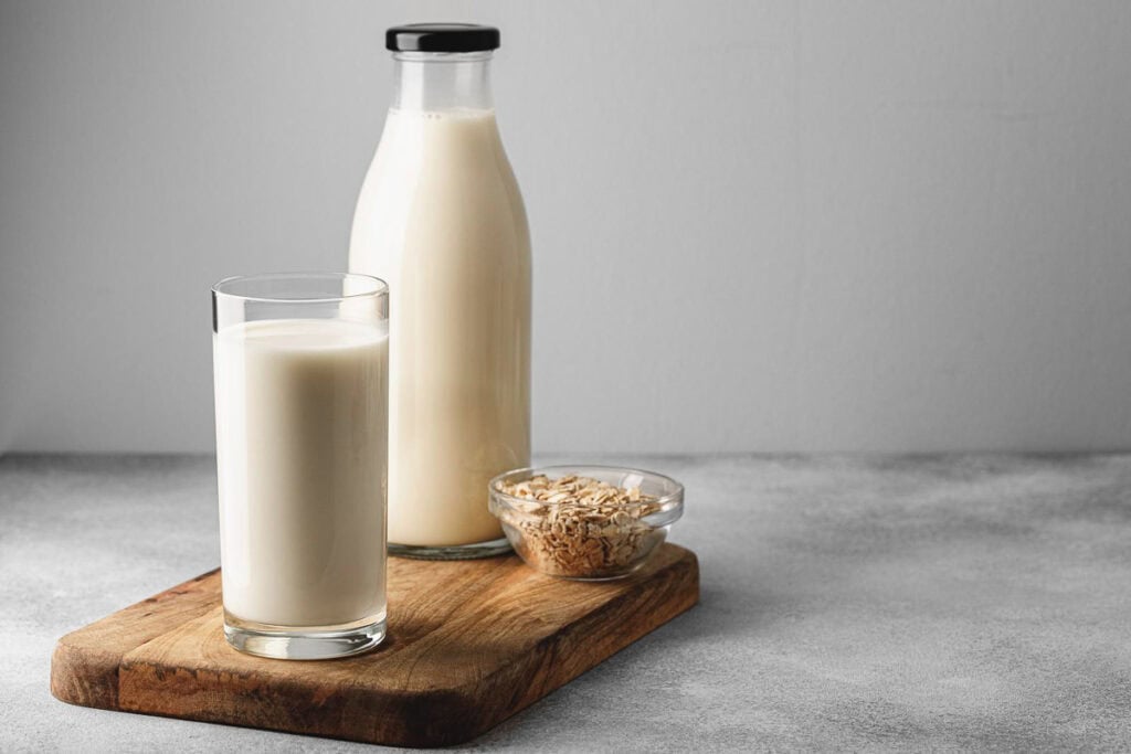 A bottle of milk, a glass of milk, and a small bowl of oats are placed on a wooden board on a gray surface.