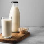 A bottle of milk, a glass of milk, and a small bowl of oats are placed on a wooden board on a gray surface.