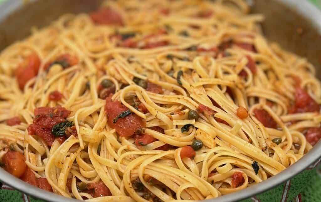A close-up of a plate of linguine pasta tossed with tomatoes, garlic, green herbs, and capers.