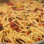 A close-up of a plate of linguine pasta tossed with tomatoes, garlic, green herbs, and capers.