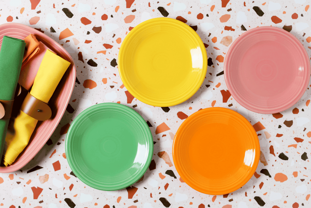 Four colorful plates in yellow, pink, green, and orange arranged on a terrazzo surface with a pink tray holding yellow and green napkins on the side.