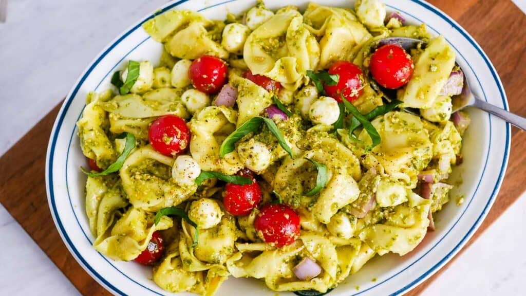 A bowl of tortellini pasta salad with pesto, cherry tomatoes, mozzarella balls, and fresh basil, served with a spoon.