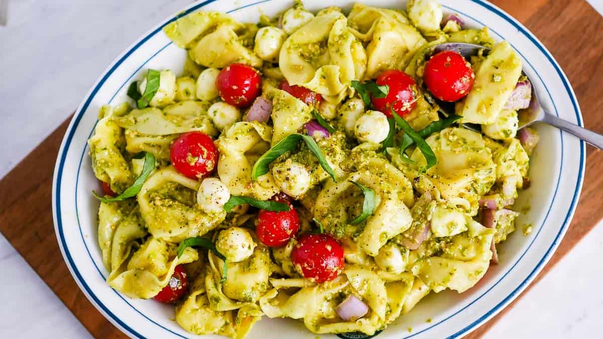A bowl of tortellini pasta salad with cherry tomatoes, mozzarella balls, basil, red onions, and a pesto dressing. A spoon is placed on the side of the dish.