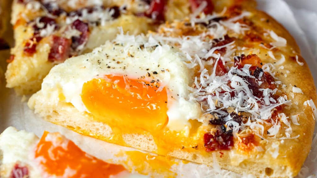 Close-up of a slice of pizza topped with a runny fried egg, grated cheese, bacon bits, and black pepper.