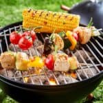 Skewers of vegetables and chunks of meat are grilling over hot coals on a barbecue, with a corn on the cob in the background.
