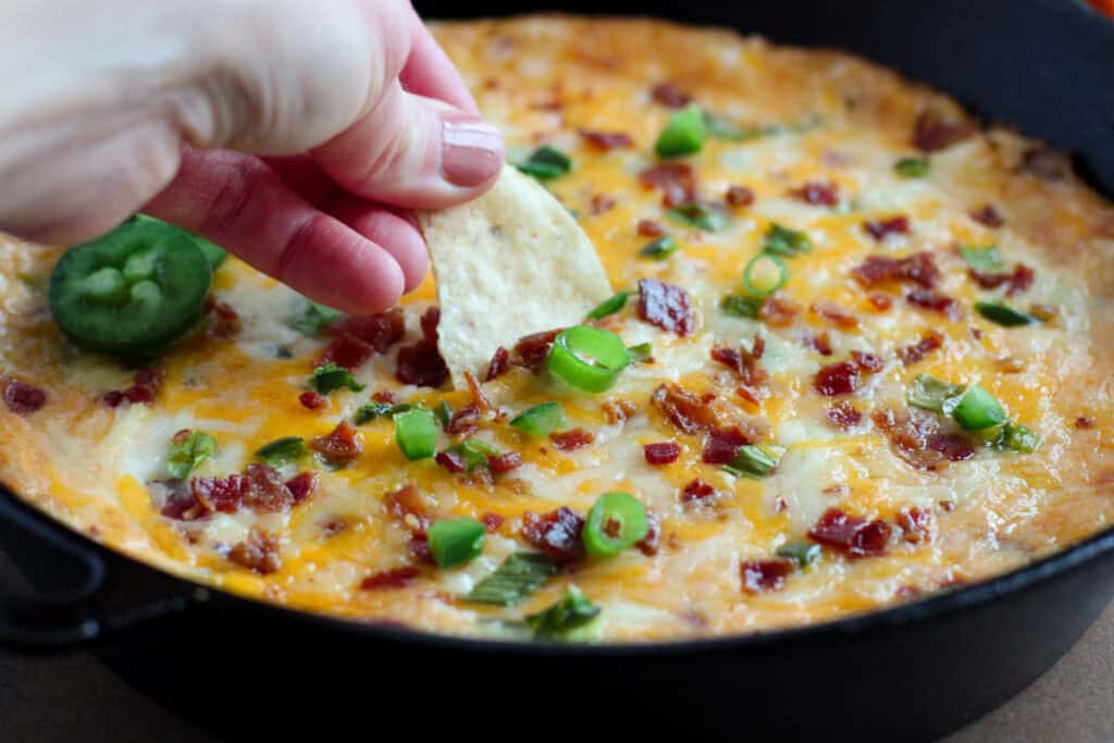 A hand dipping a chip into a skillet containing a melted cheese dish topped with chopped green onions, bacon bits, and a slice of jalapeño.