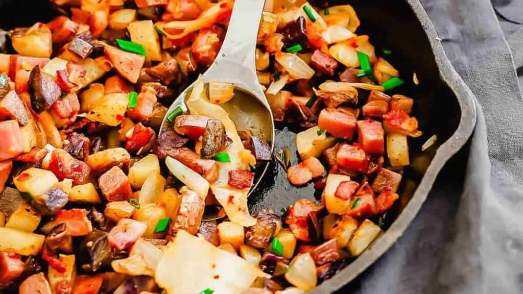 A cast-iron skillet filled with a sautéed mixture of diced potatoes, onions, and chunks of meat, garnished with chopped herbs.