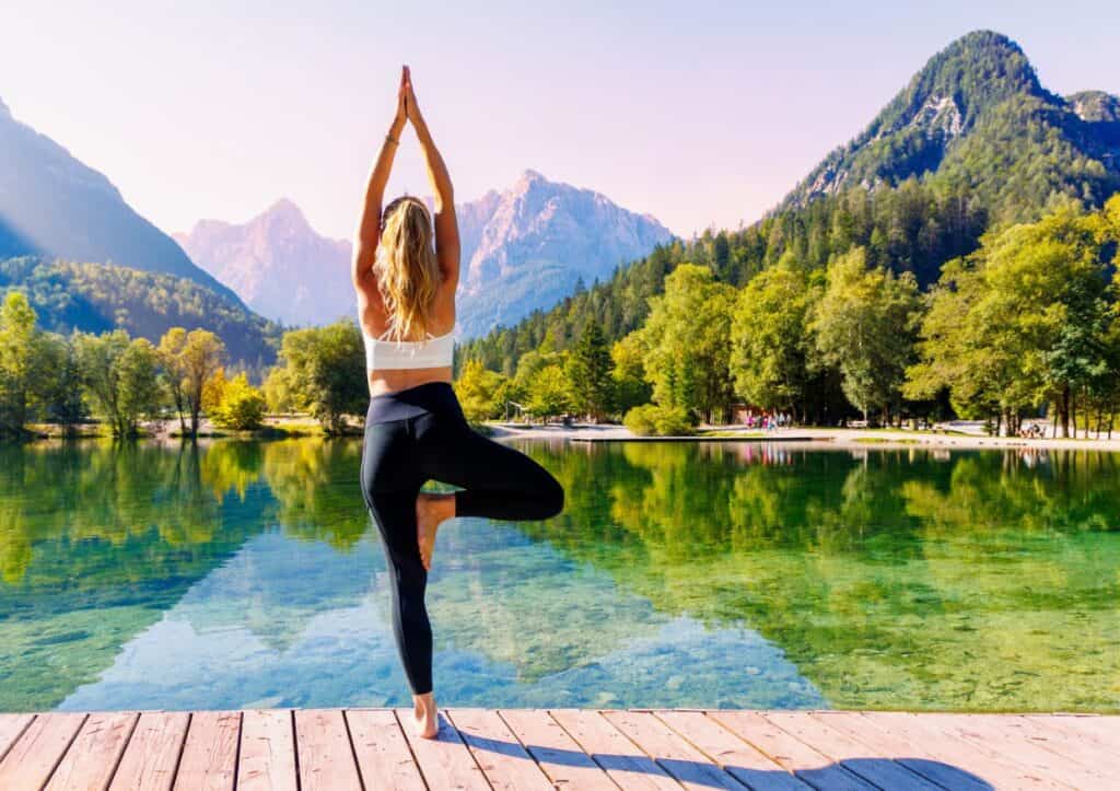 A person stands on a wooden dock in a yoga pose, overlooking a clear lake with reflections of trees and mountains under a sunny sky.