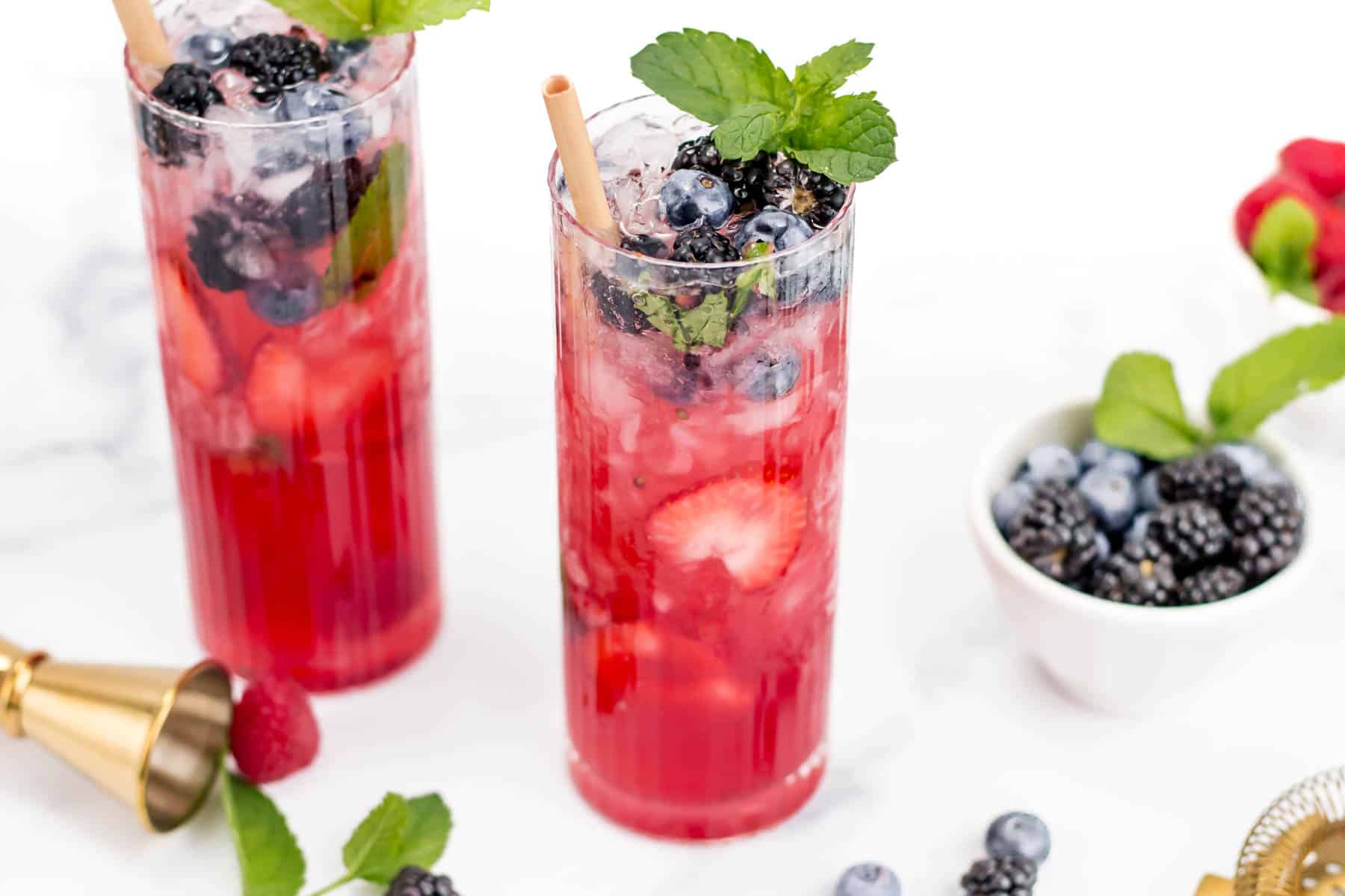 Two tall glasses of berry-infused red drink garnished with mint leaves and fresh berries. A bowl of berries, a measuring jigger, and loose berries are placed around the glasses on a white surface.
