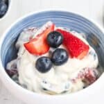 A bowl of creamy berry salad topped with fresh blueberries and sliced strawberries. A gold spoon is placed beside the bowl, and there is a small dish of blueberries in the background.