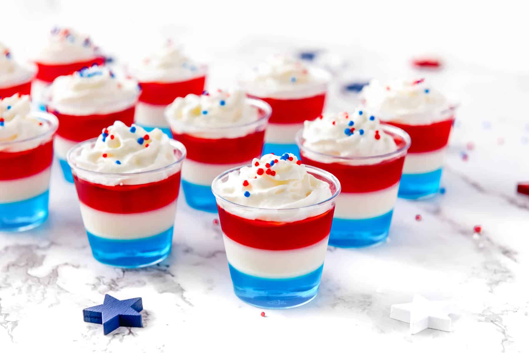 Several cups of layered red, white, and blue gelatin topped with whipped cream and sprinkles are arranged on a white surface.