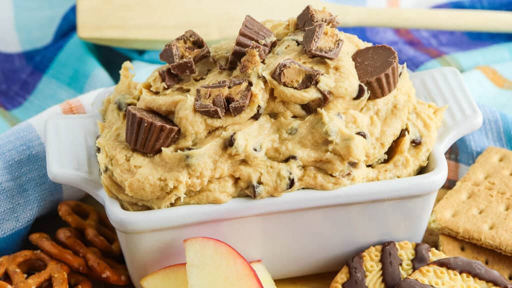 A white dish filled with cookie dough dip topped with chopped peanut butter cups. Surrounding the dish are pretzels, an apple slice, graham crackers, and a chocolate-covered cookie.