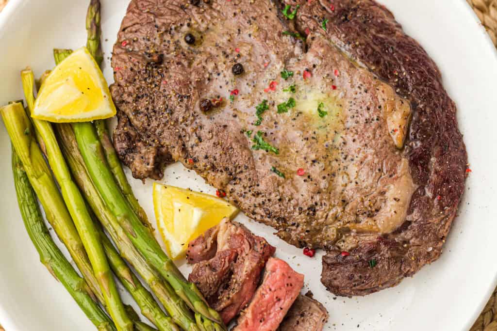 A cooked steak with grilled asparagus and lemon wedges is served on a white plate. A wedge of steak and some parsley are on top of the main steak.