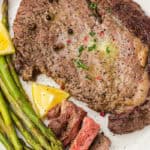 A cooked steak with grilled asparagus and lemon wedges is served on a white plate. A wedge of steak and some parsley are on top of the main steak.