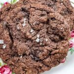 A close-up of a large chocolate cookie sprinkled with coarse salt, placed on a decorative plate with pink rose patterns.