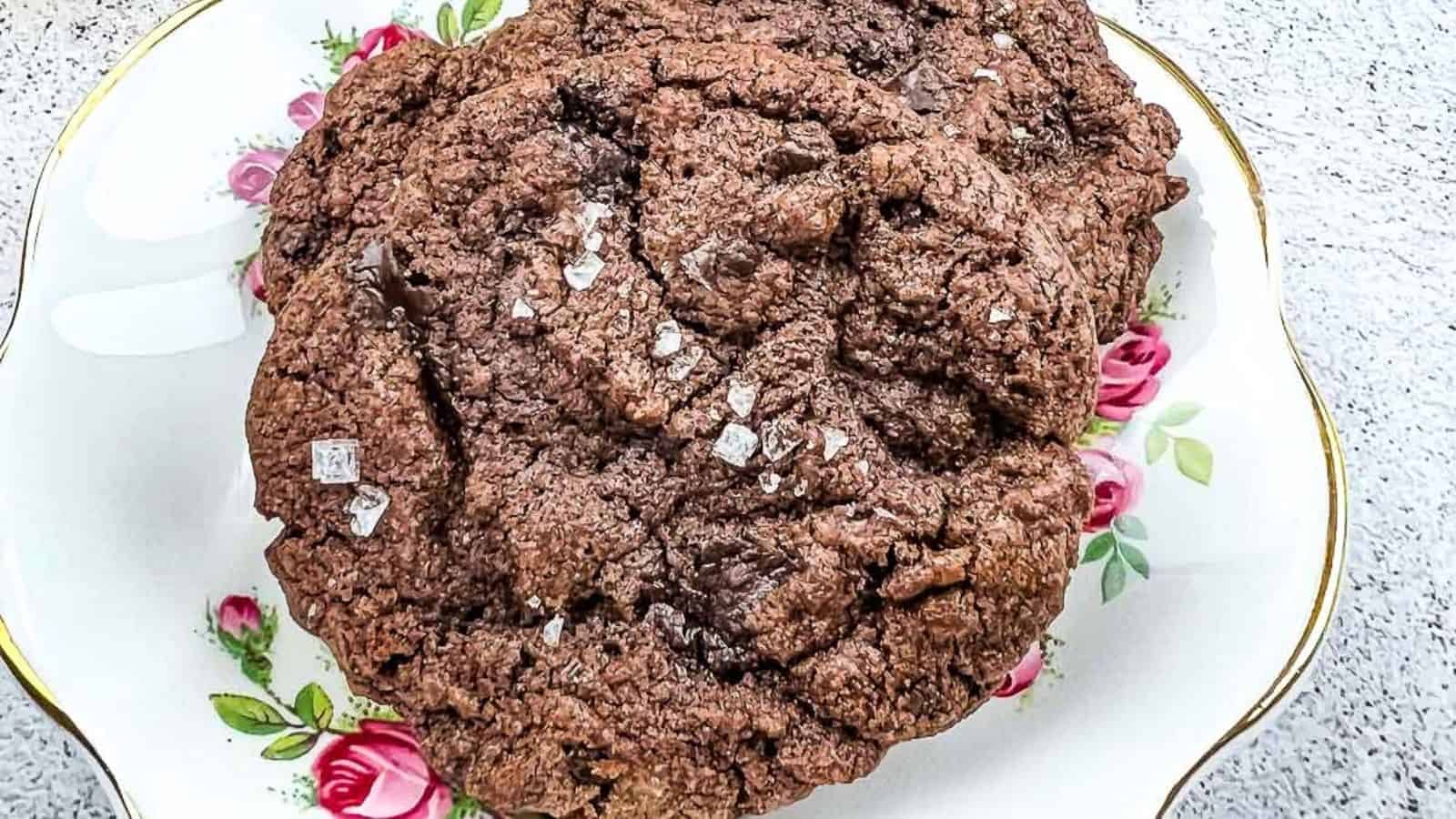 A close-up of a large chocolate cookie sprinkled with coarse salt, placed on a decorative plate with pink rose patterns.