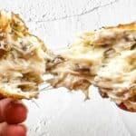 A close-up shot of hands pulling apart a crispy, golden-brown, cheese-filled snack with a gooey, melted cheese center against a textured white background.