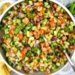 A bowl of chopped vegetable salad with cucumbers, tomatoes, red onions, and parsley, next to a yellow checkered cloth and fresh parsley.