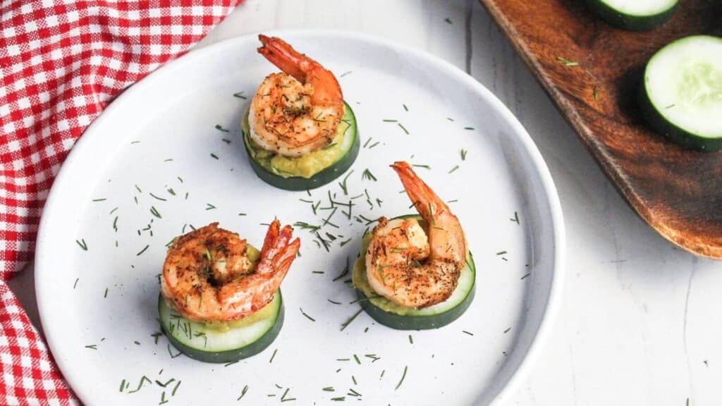 Three grilled shrimp on cucumber slices, garnished with dill on a white plate next to a red checkered napkin.