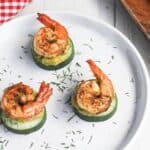 Three grilled shrimp on cucumber slices, garnished with dill on a white plate next to a red checkered napkin.