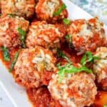 A white rectangular plate features meatballs topped with grated cheese and garnished with chopped herbs, resting on a bed of tomato sauce.