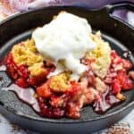 A cast iron skillet with strawberry rhubarb cobbler topped with a scoop of vanilla ice cream on a wooden surface with a cloth napkin beside it.