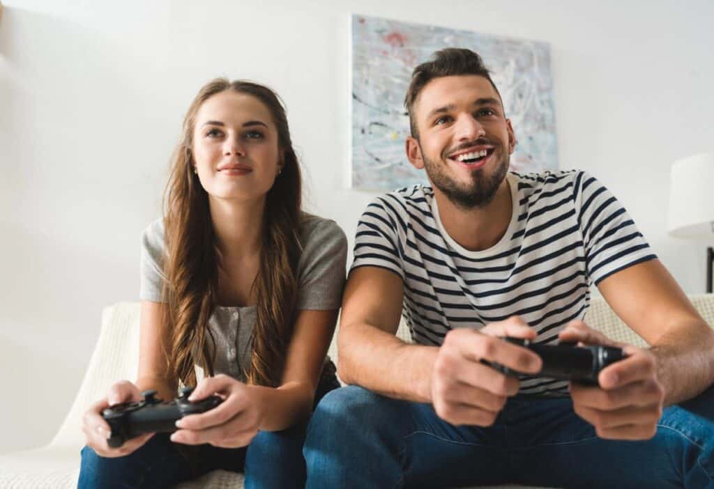A smiling man and woman sit on a couch, each holding video game controllers, fully immersed in playing stress-relieving games.
