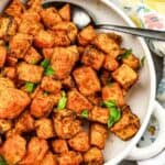 A white bowl filled with seasoned roasted sweet potato cubes, garnished with chopped herbs, with a metal serving spoon on the side and a printed napkin partially visible.