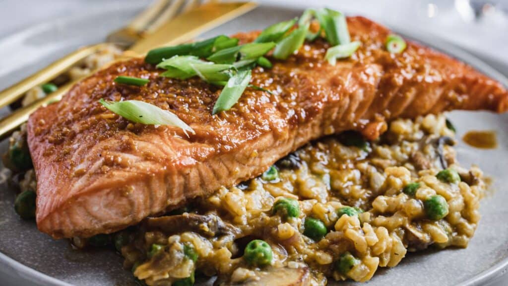 A plate of cooked salmon topped with green onions, served over a bed of risotto mixed with peas and mushrooms, with a fork and knife placed on the side.