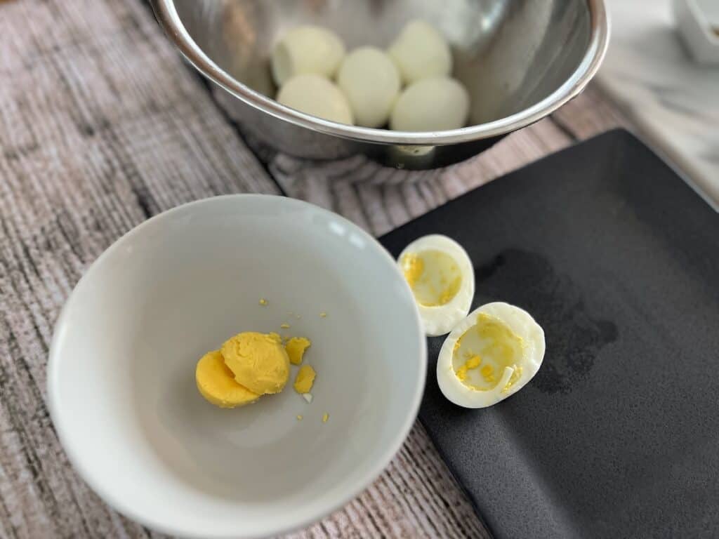 A hard-boiled egg is cut in half with the yolk placed in a separate white bowl. There is a metal bowl with whole hard-boiled eggs in the background and a black plate beside the bowls.