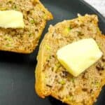 A sliced muffin with visible nuts and green specks is topped with butter and placed on a black plate.