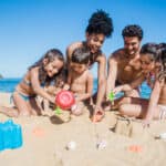 A Caribbean family enjoys fun in the sun as children and two adults build sandcastles together on a sandy beach, with the blue ocean shimmering in the background.