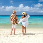 A man and woman holding hands, standing on a sandy beach of one of the Caribbean's Most Enchanting Islands. Both are wearing hats and summer clothing, facing the turquoise ocean with a partly cloudy sky above.