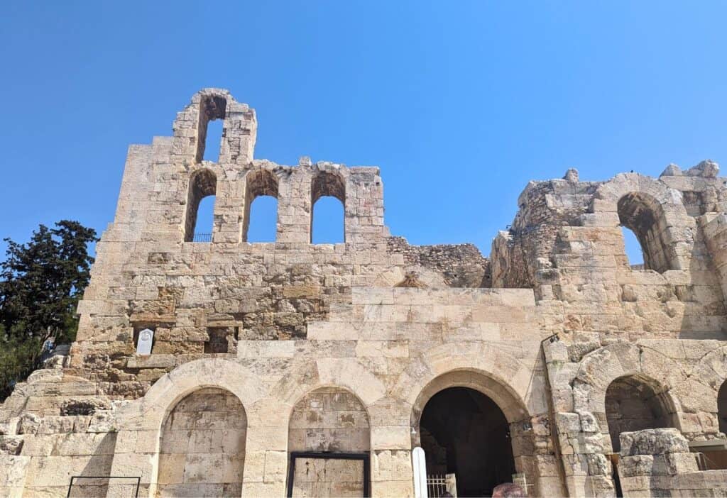 The image shows the ruins of an ancient stone building with multiple arched windows and doorways under a clear blue sky.