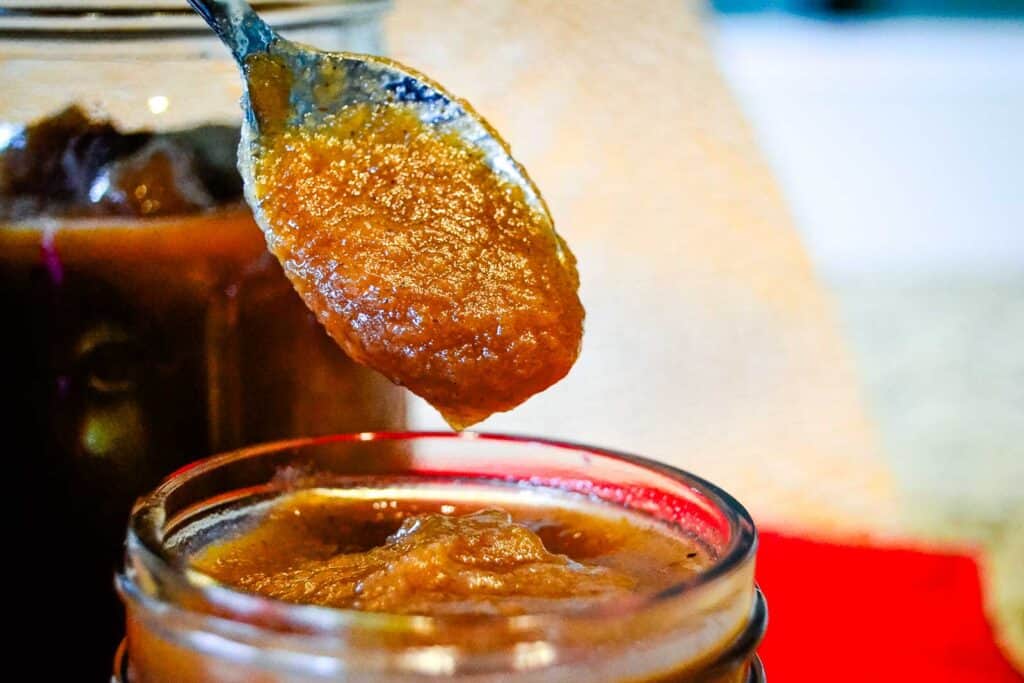 A spoonful of apple butter is being held over an open jar, with another jar in the background.