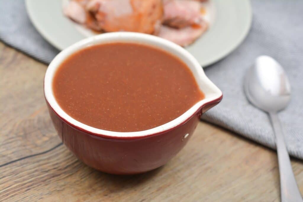 A red ceramic bowl filled with brown sauce sits on a wooden table next to a spoon and a plate with sliced meat.