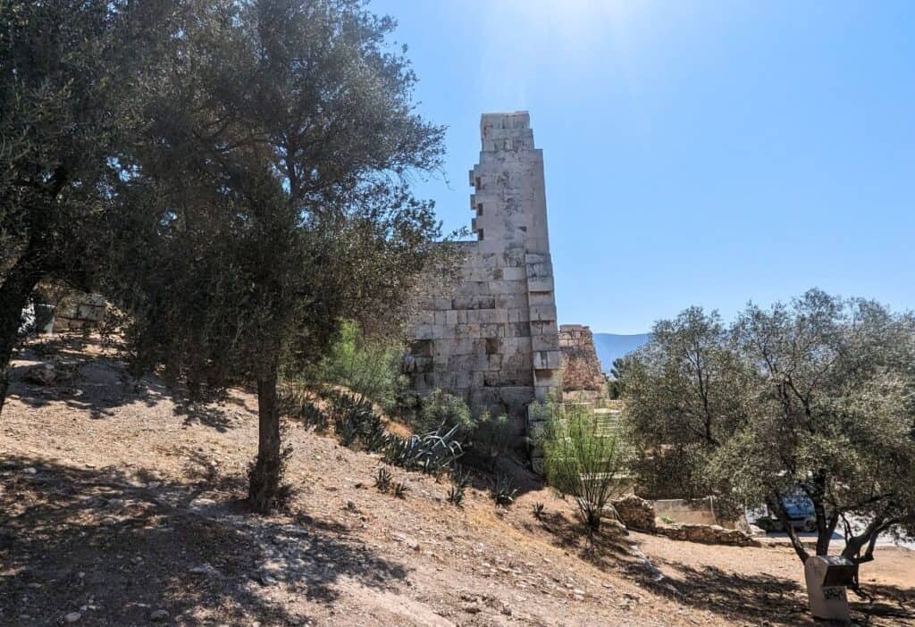Ancient stone tower ruins stand under a clear blue sky, surrounded by olive trees and dry, rocky terrain.