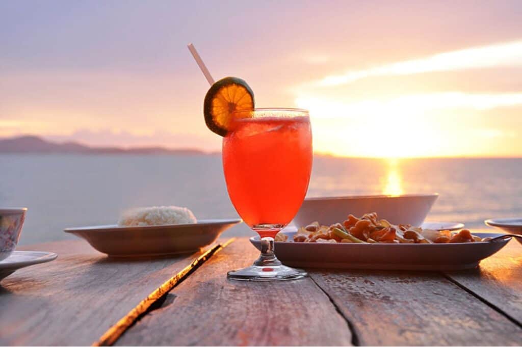 A glass of red-orange drink with a straw and lime slice on a wooden table, surrounded by plates of food, against a sunset backdrop over the water.