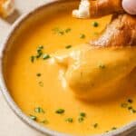 A hand dips a piece of bread into a bowl of creamy orange-yellow soup or sauce, garnished with chopped green herbs.