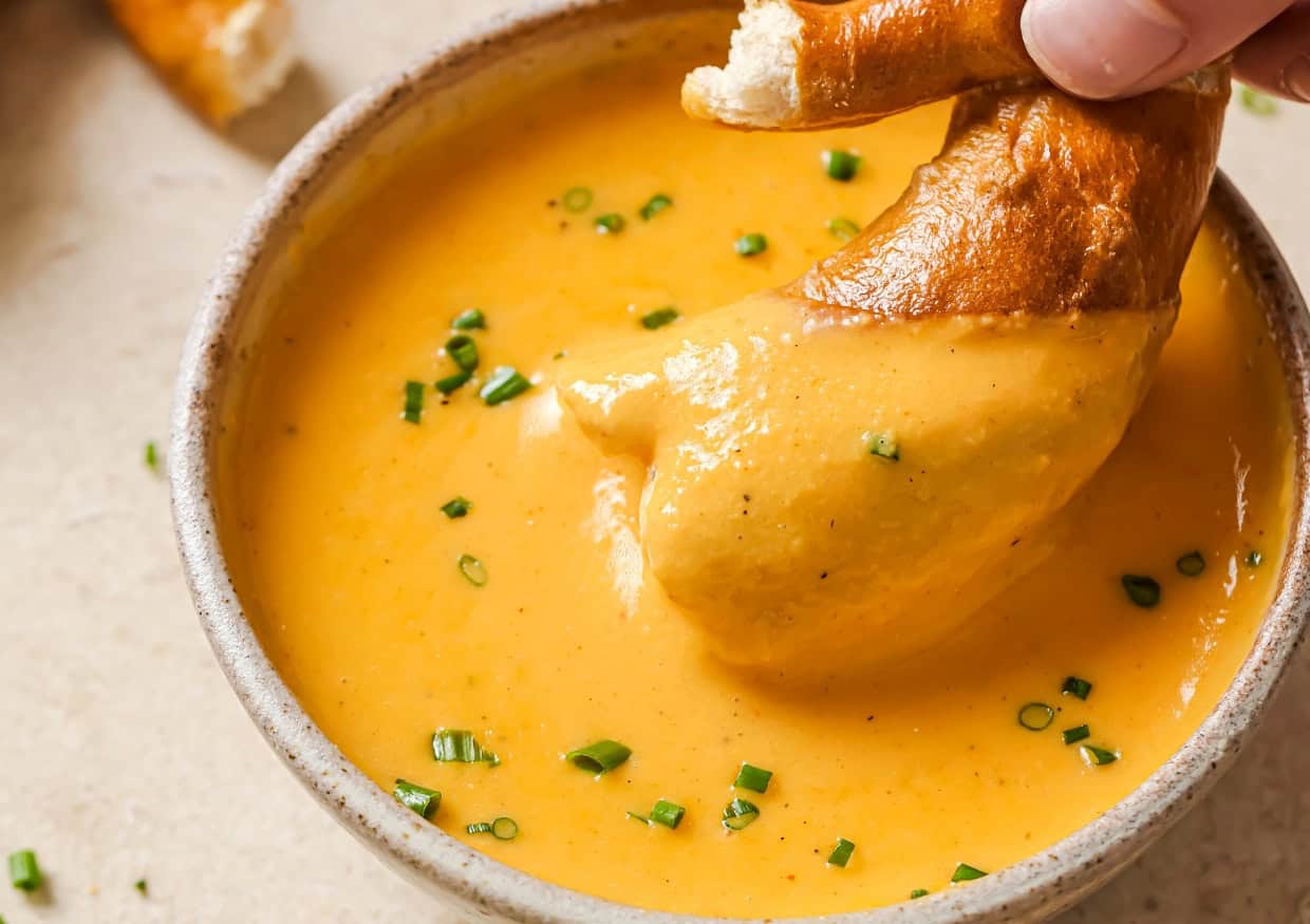 A piece of pretzel being dipped into a bowl of cheese sauce garnished with chopped chives.