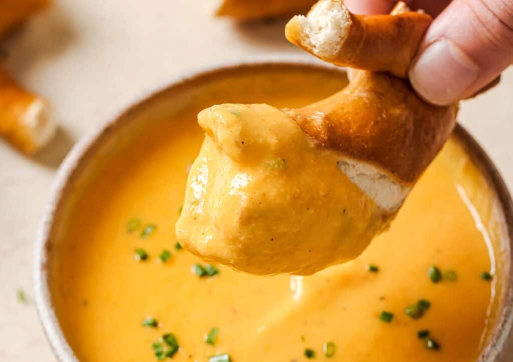 A piece of pretzel being dipped into a bowl of cheese sauce garnished with chopped chives.