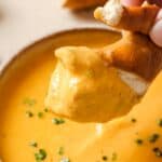 A piece of pretzel being dipped into a bowl of cheese sauce garnished with chopped chives.