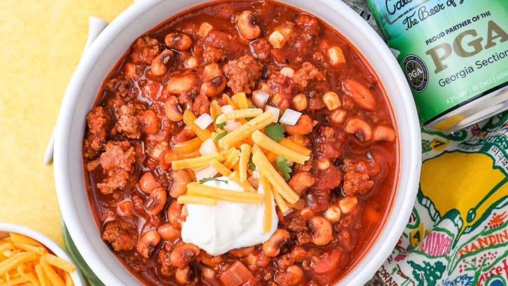 A bowl of hearty chili topped with shredded cheese and a dollop of sour cream. The dish contains various beans, ground beef, and vegetables. A can and a bowl of shredded cheese are beside it.