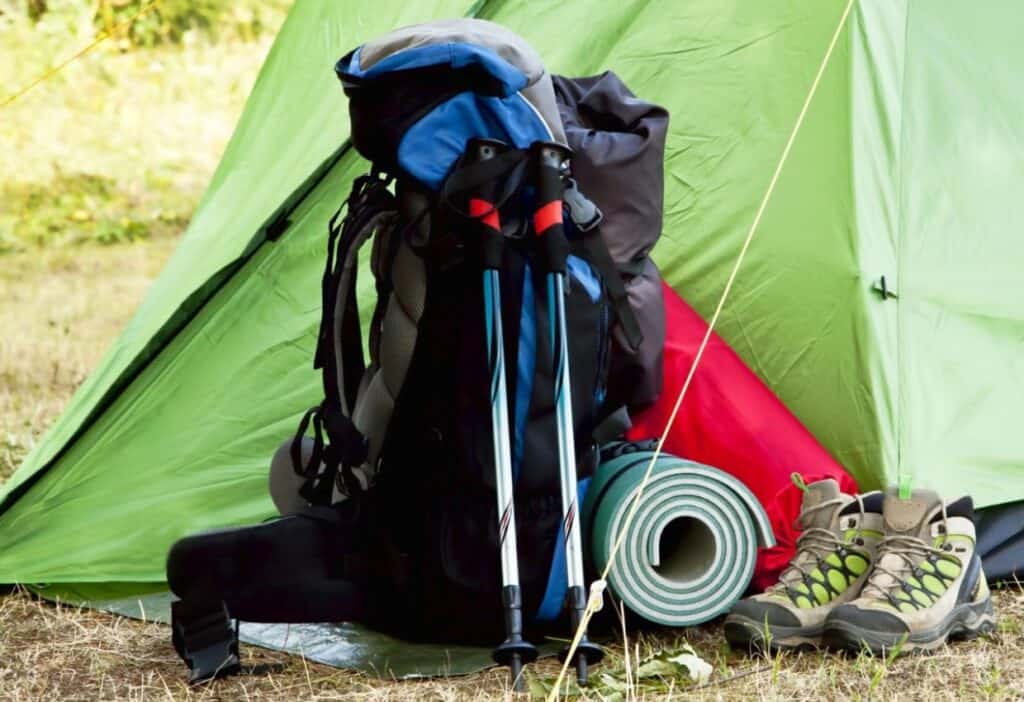 A camping backpack, trekking poles, rolled-up sleeping pad, and hiking boots are positioned in front of a green and red tent set up outdoors.